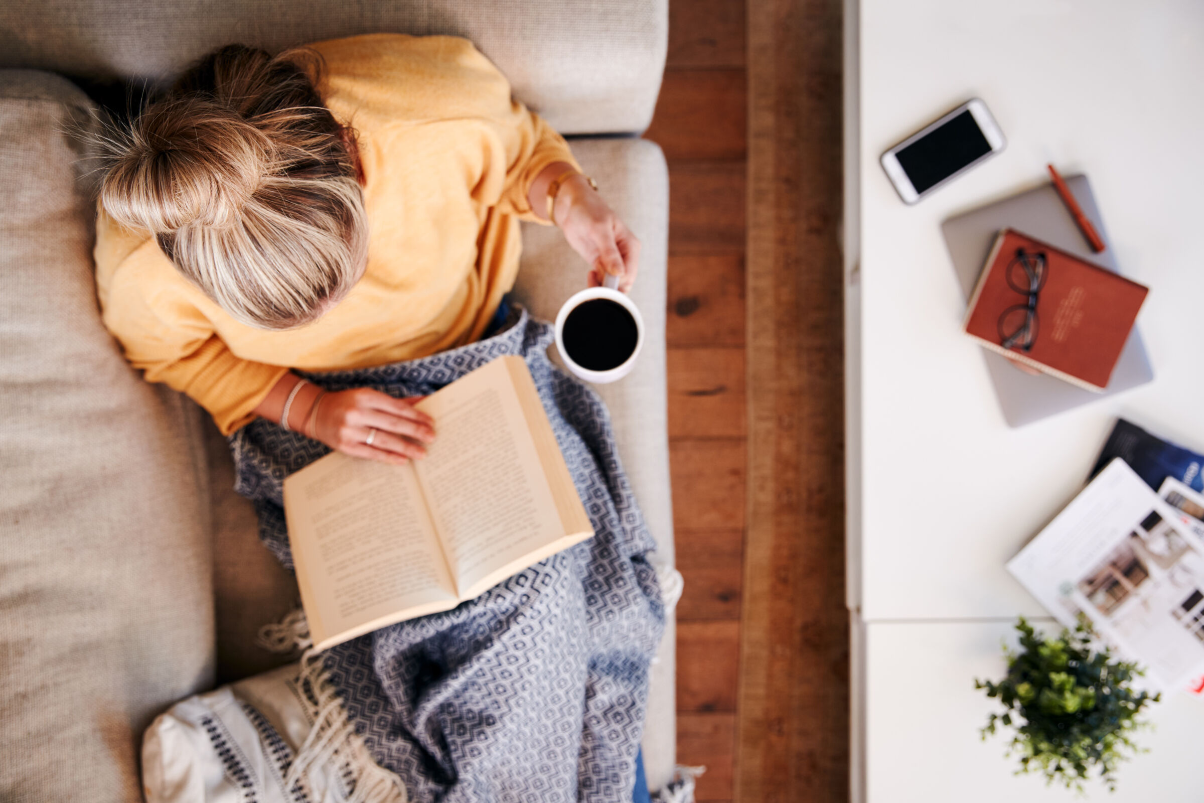 High angle view of woman reading on couch and drinking coffee
