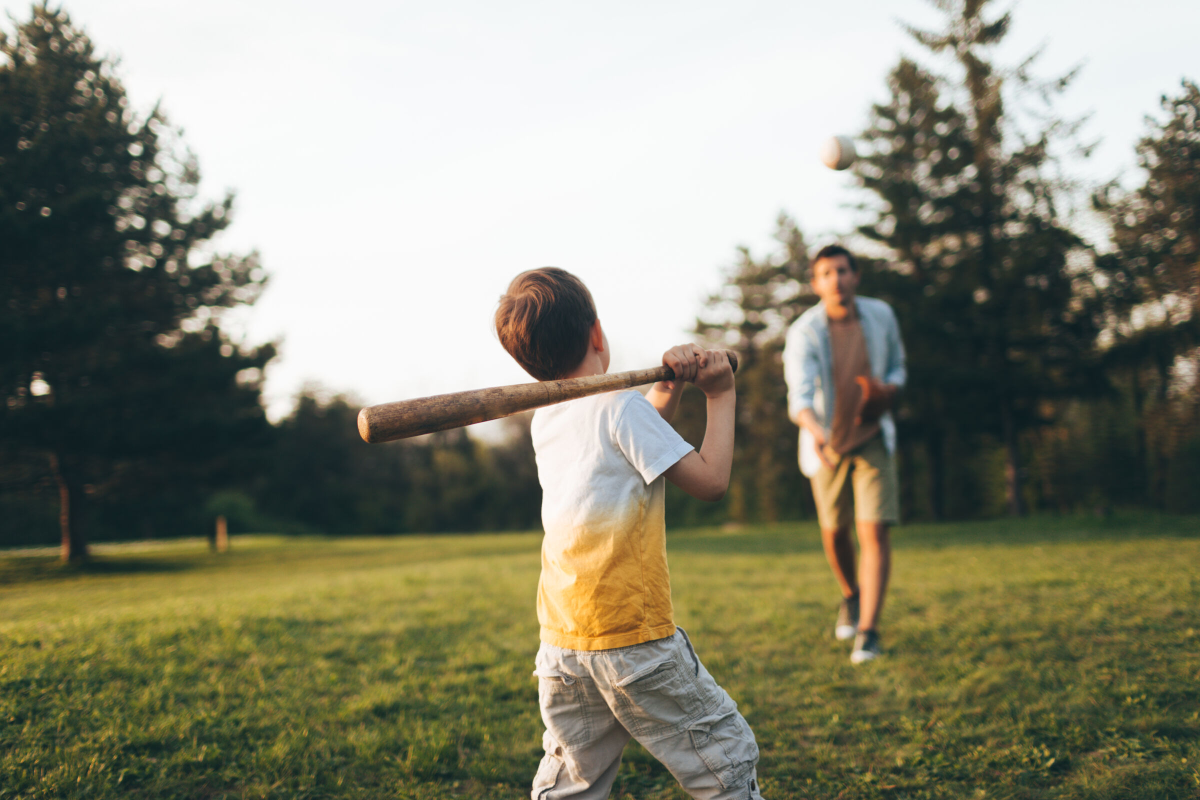 Closeup of a boy swinging a baseball bat with his dad tossing a pitch on a sunny day.