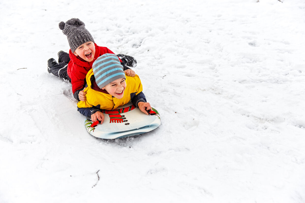 Two boys happily slide down on a sled in the winter with snow covering the ground.